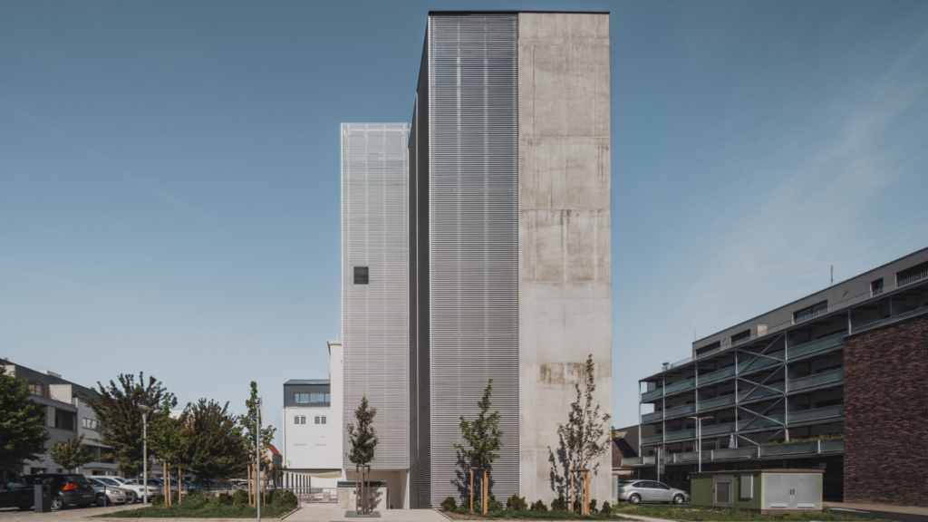 Two tall concrete towers with metal louvers rise above a small urban plaza under a clear blue sky, surrounded by parked cars and trees.