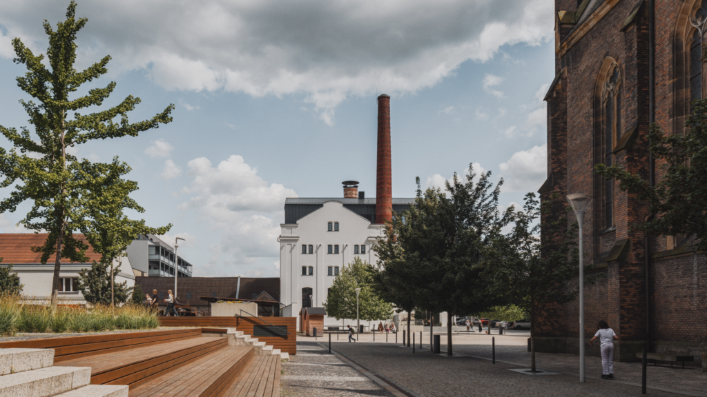 A street scene with a tall red brick chimney and white building, trees lining the plaza, and a church-like brick facade on the right under a cloudy sky.
